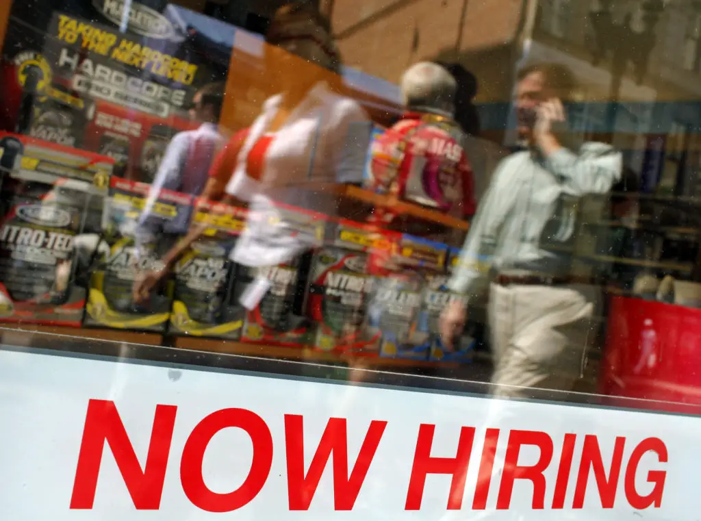 Pedestrians walk past a "Now Hiring" sign in the window of a GNC shop in Boston