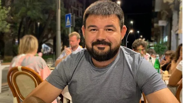 Man wearing grey shirt in sitting in outdoor dining aera smiles at camera.